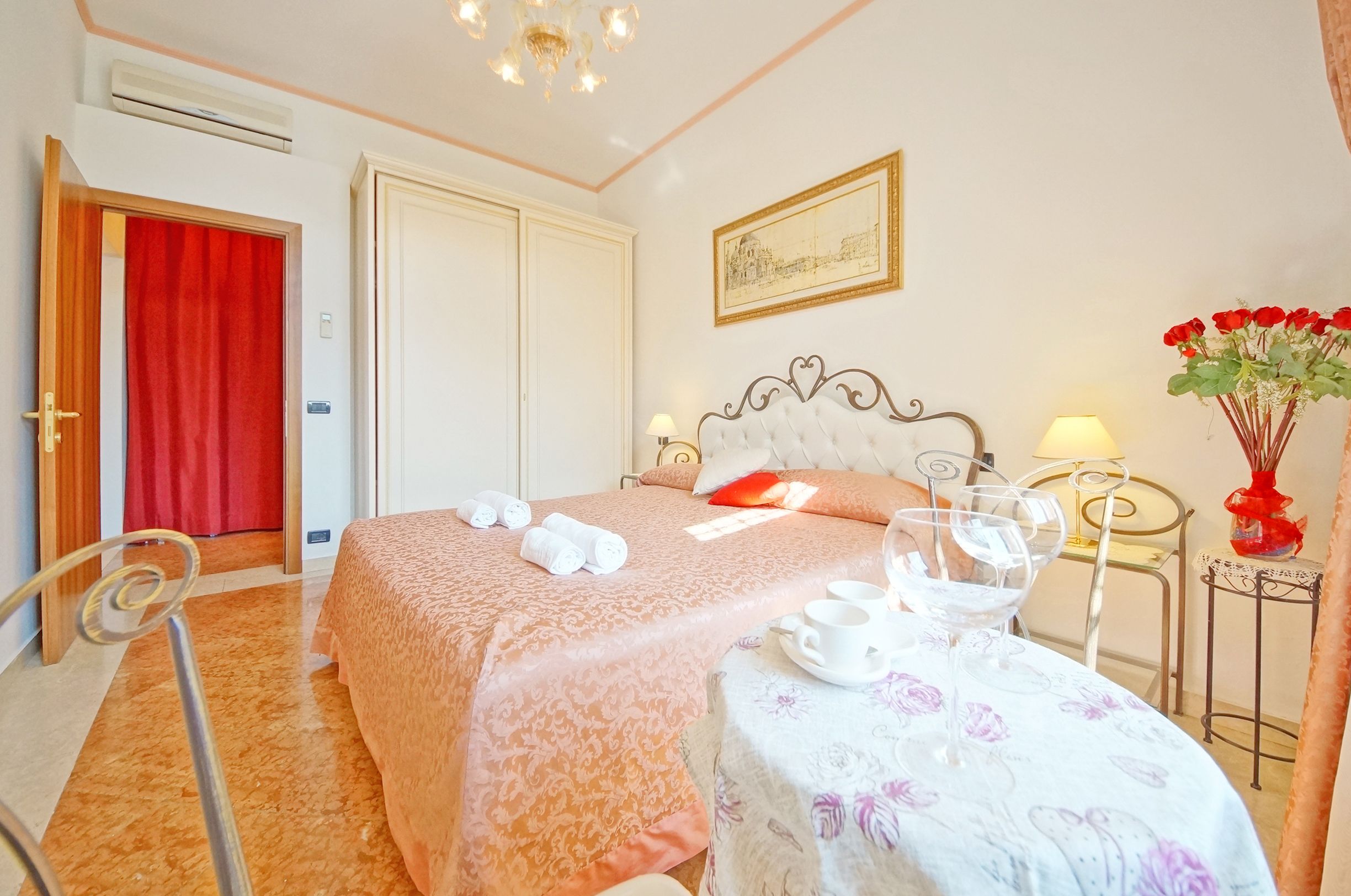 Een hotelkamer met een bed, een tafel met een tafelkleed met bloemen, een vaas met rode bloemen, en een schilderij aan de muur.