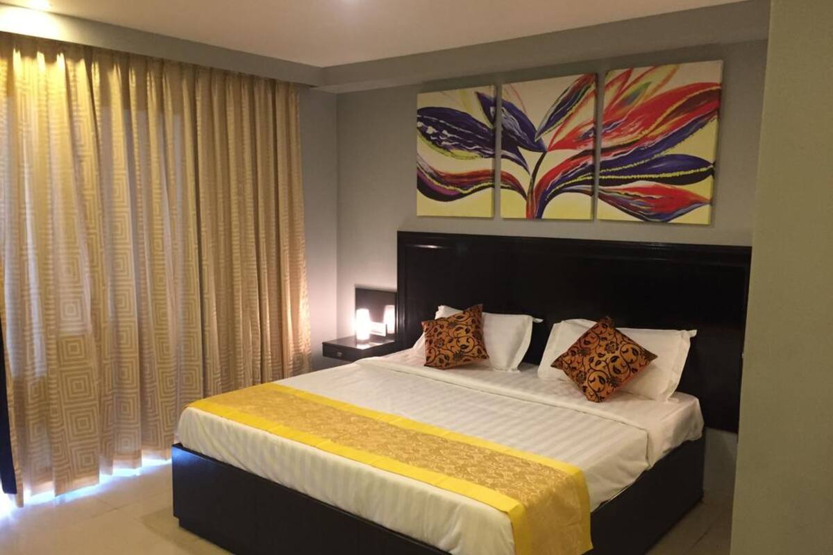 Habitación de hotel con una cama grande, cabecera, mesita de noche, lámpara, ventana con cortinas y un cuadro abstracto y colorido en la pared.