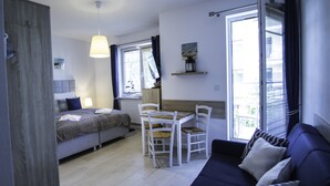 Een moderne slaapkamer met een bed, een bureau en een bank. Er is een raam met zonwering, een hanglamp en een klein plankje met decoratieve objecten.