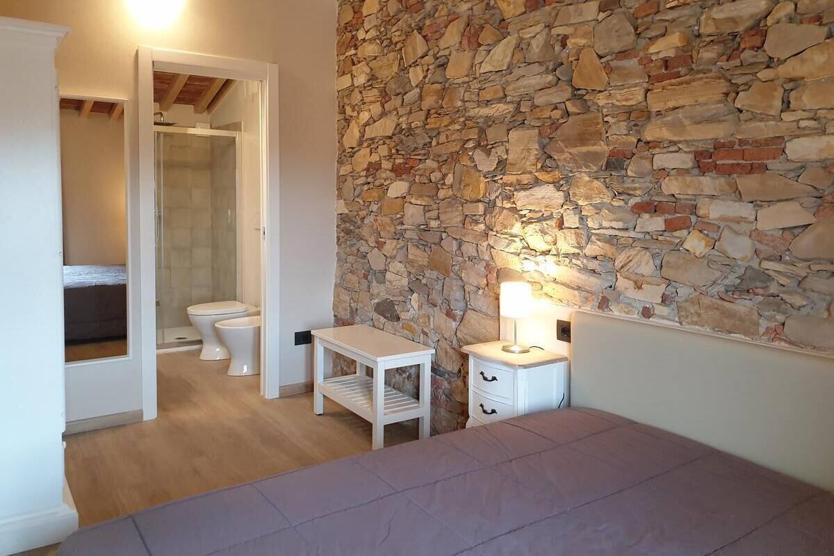 A bedroom with a stone wall, a bed, a bedside table with a lamp, and a bathroom with a toilet and shower.