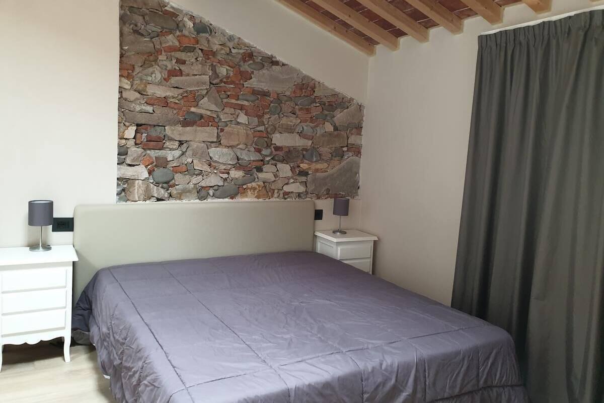 A bedroom with a stone wall, a bed with a grey bedspread, a nightstand with a lamp, and a window with a grey curtain.