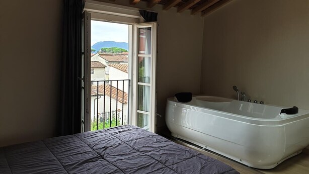 A bedroom with a large bed, a bathtub, and a balcony with a view of buildings and mountains. - B&B Il Tiglio (Lucca)