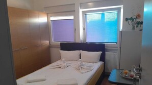 A single bed with white linens, a wooden wardrobe, a small table with a tray of items, and a window with blinds. - Pension und Monteurzimmer VICTORIA (Ludwigshafen)