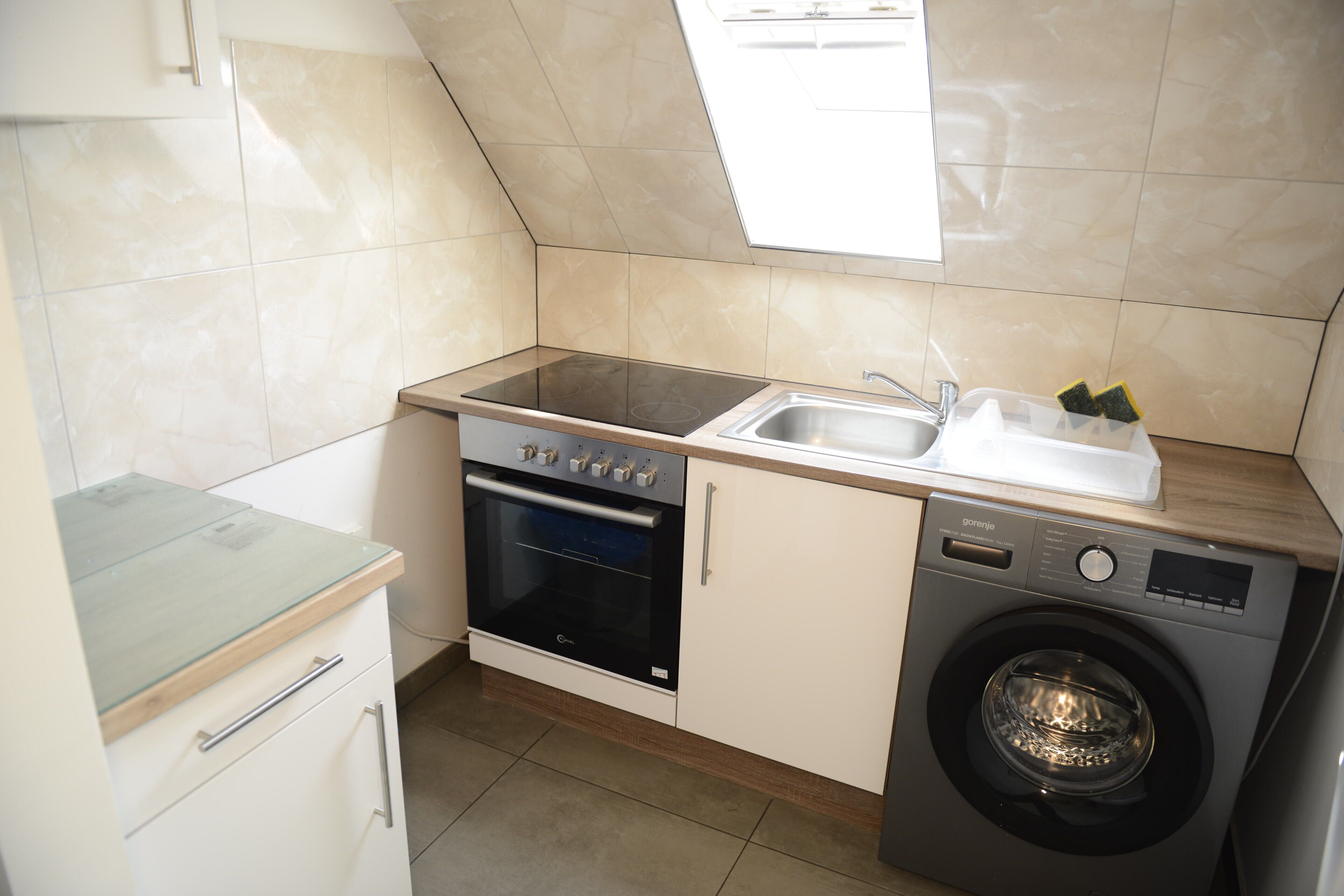 A compact kitchen with a built-in oven, a washing machine, and a window above the sink.