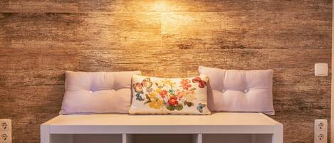 A white sofa with a floral pillow against a wooden wall, a small table with a lamp, and a power outlet on the wall.