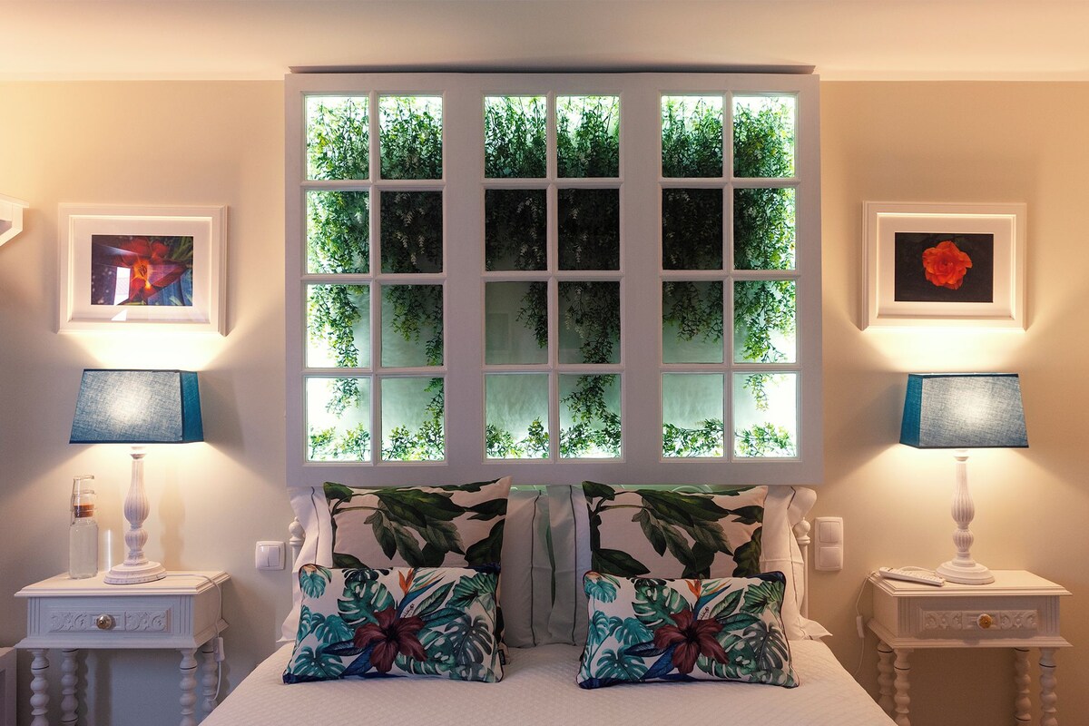 Une chambre avec une grande fenêtre, deux tables de chevet avec des lampes et des coussins à motifs floraux.