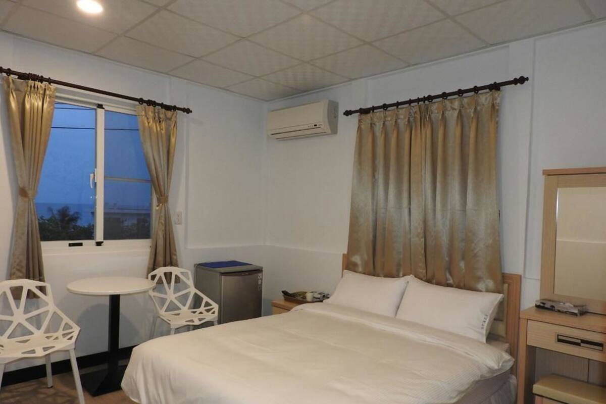 A hotel room with a bed, a desk, a chair, a small table, and a window with curtains.