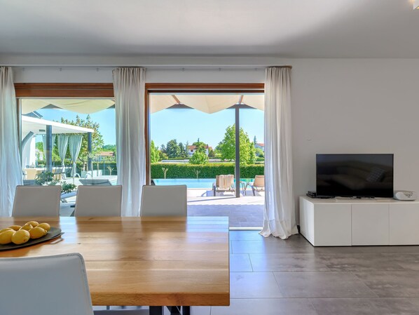 Relax in this spacious living room with a large TV, dining table, and pool views through sheer curtains.