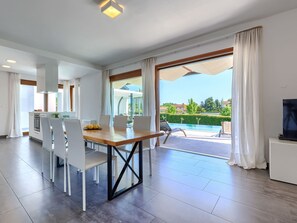 Una cocina moderna con gabinetes blancos y un horno empotrado, un comedor con mesa de madera y sillas blancas, y un amplio ventanal corredero que da a una zona de piscina.