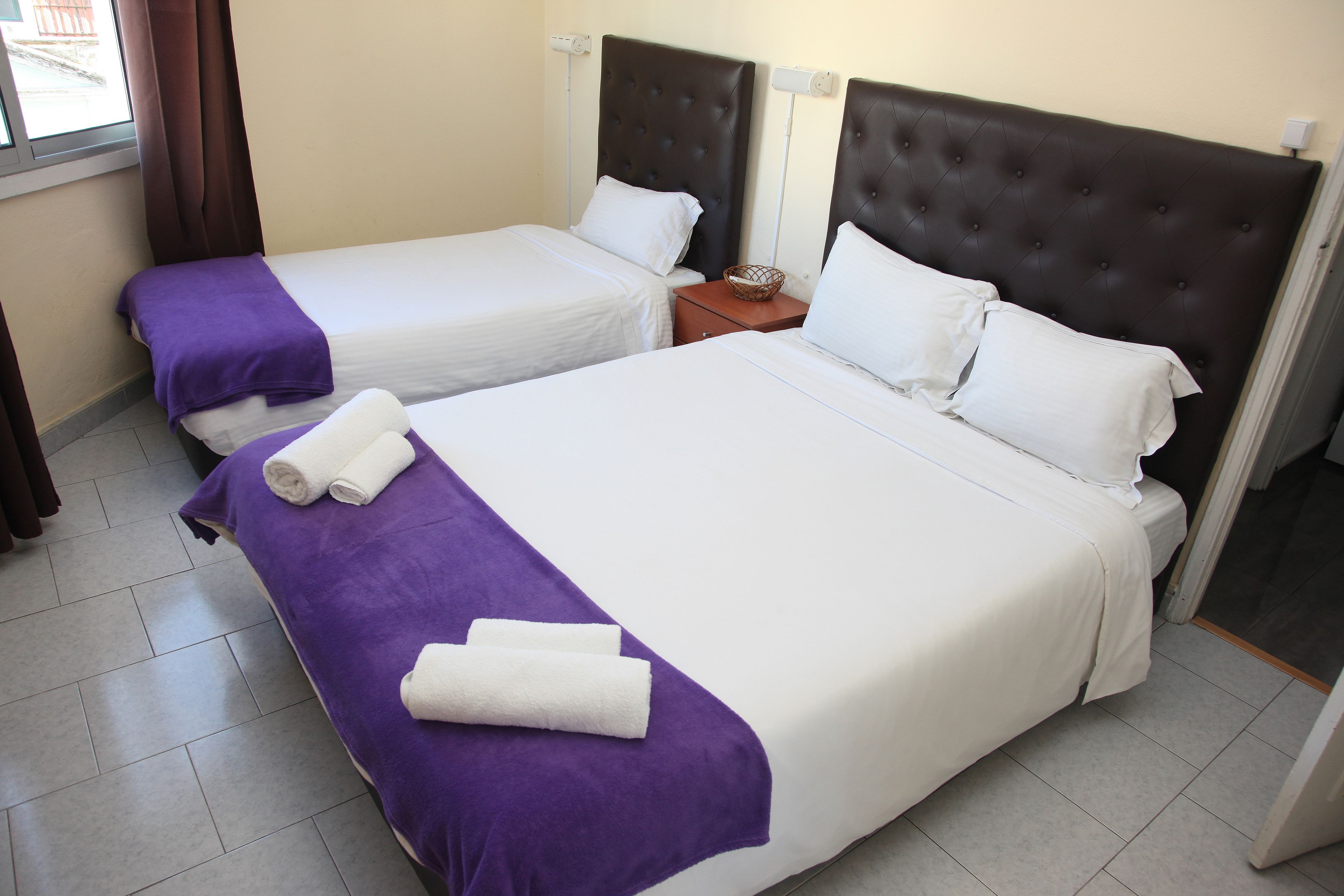 A hotel room with two beds, each with a headboard, a nightstand, and a window with curtains.