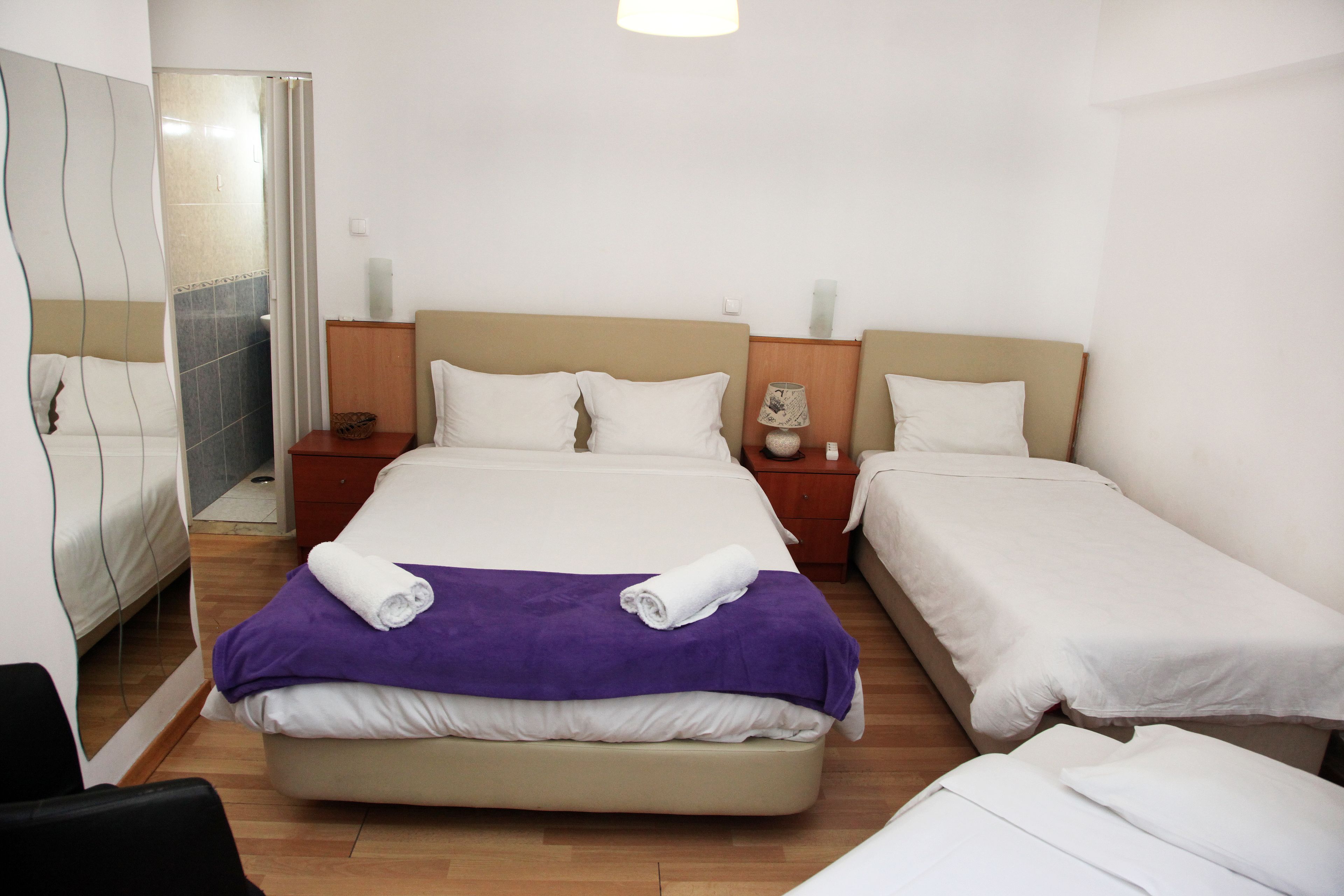 Habitación de hotel con una cama grande, una cama más pequeña, una silla, una mesita de noche, un espejo y un baño con ducha.