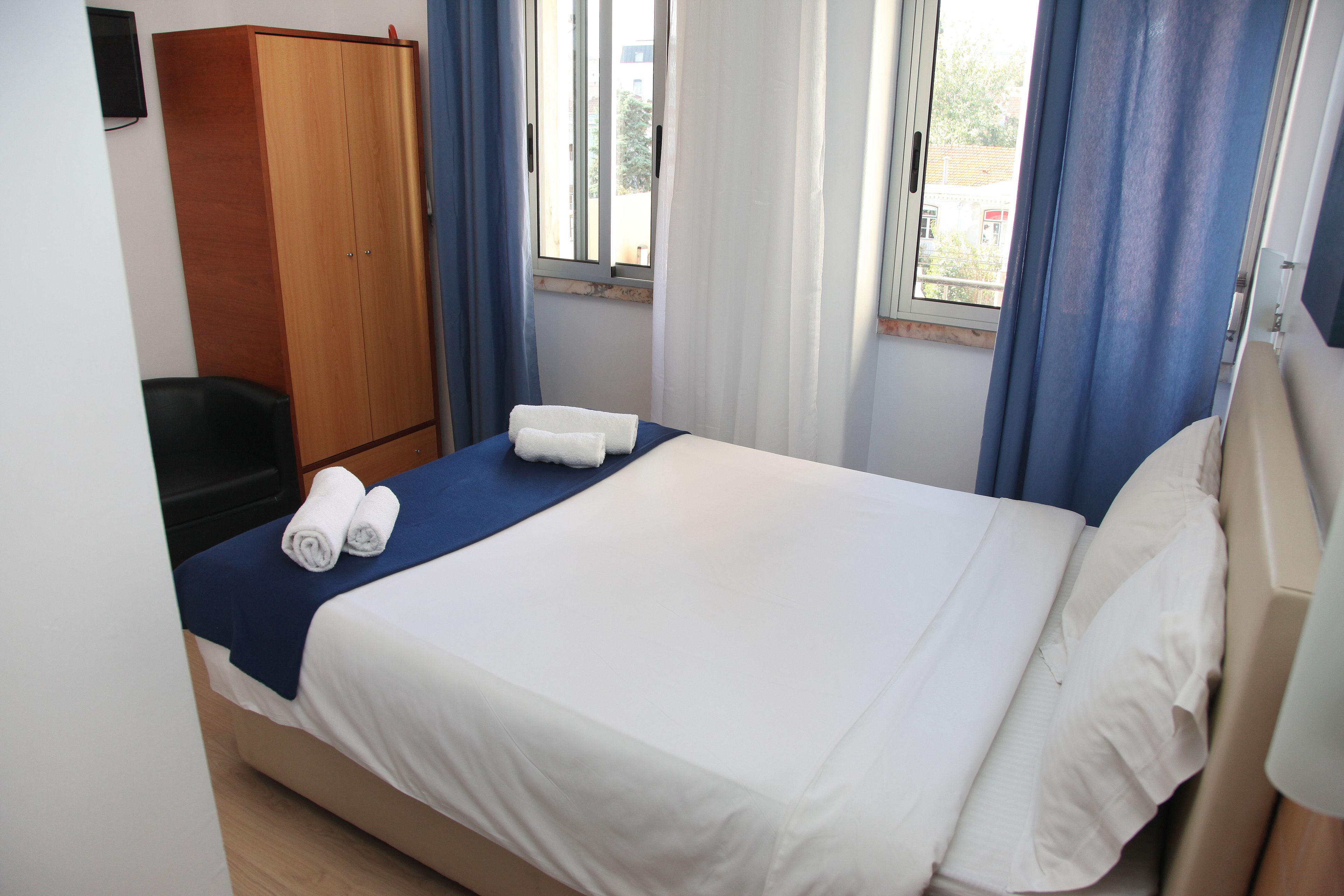 Una cama bien hecha con sábanas blancas y una colcha azul, un armario de madera y una ventana con cortinas azules.