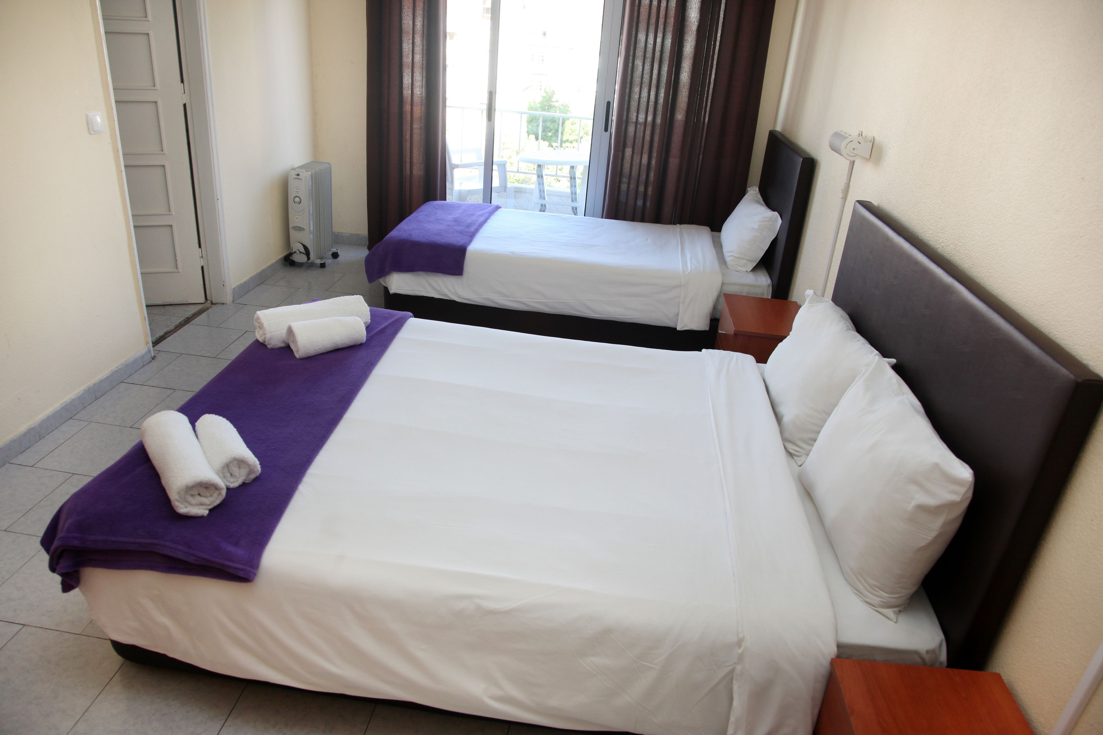 A hotel room with two beds, each with white linens and purple towels. There is a small table and a power outlet on the wall.
