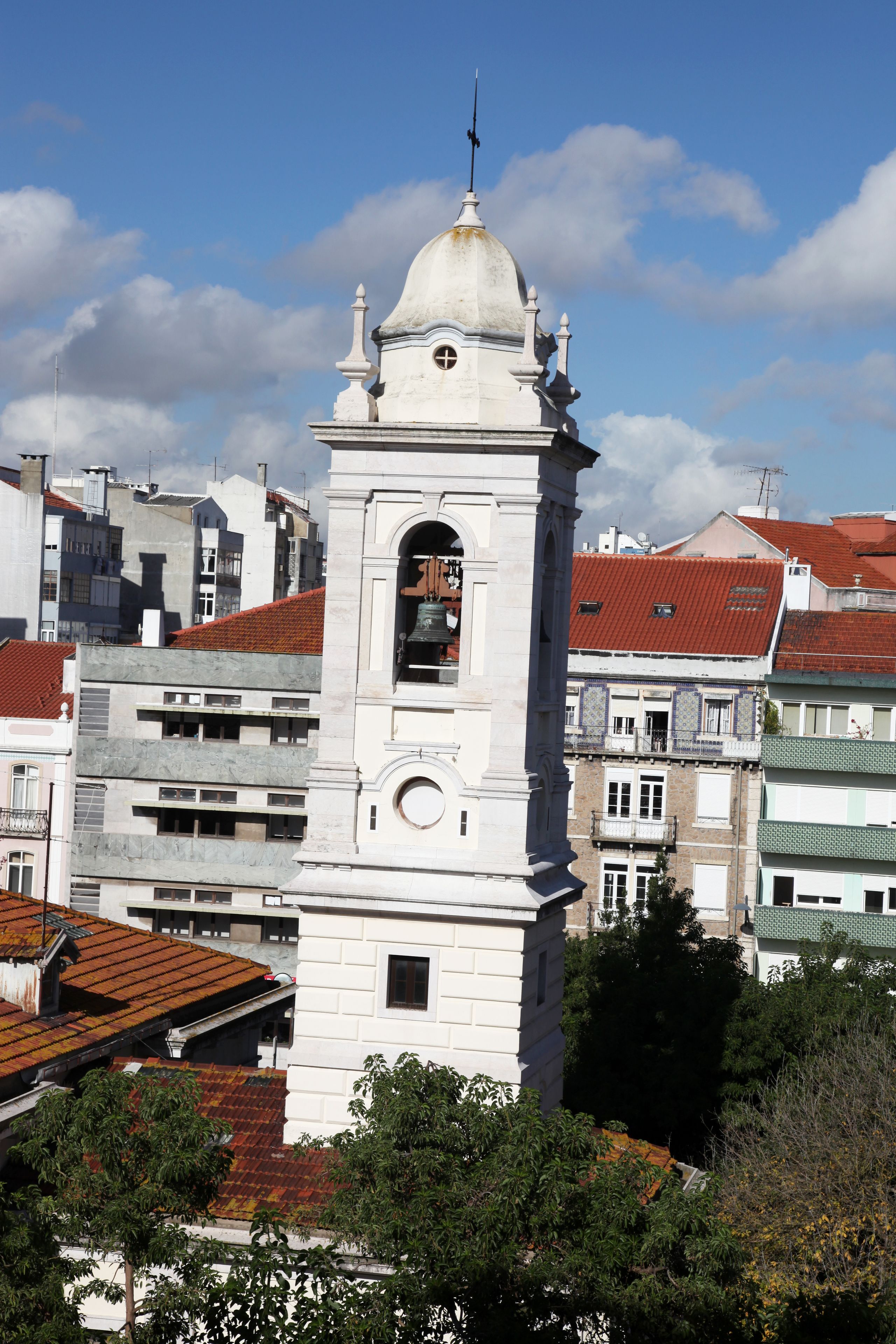 A white clock tower with a dome and bell, surrounded by buildings with red-tiled roofs.
