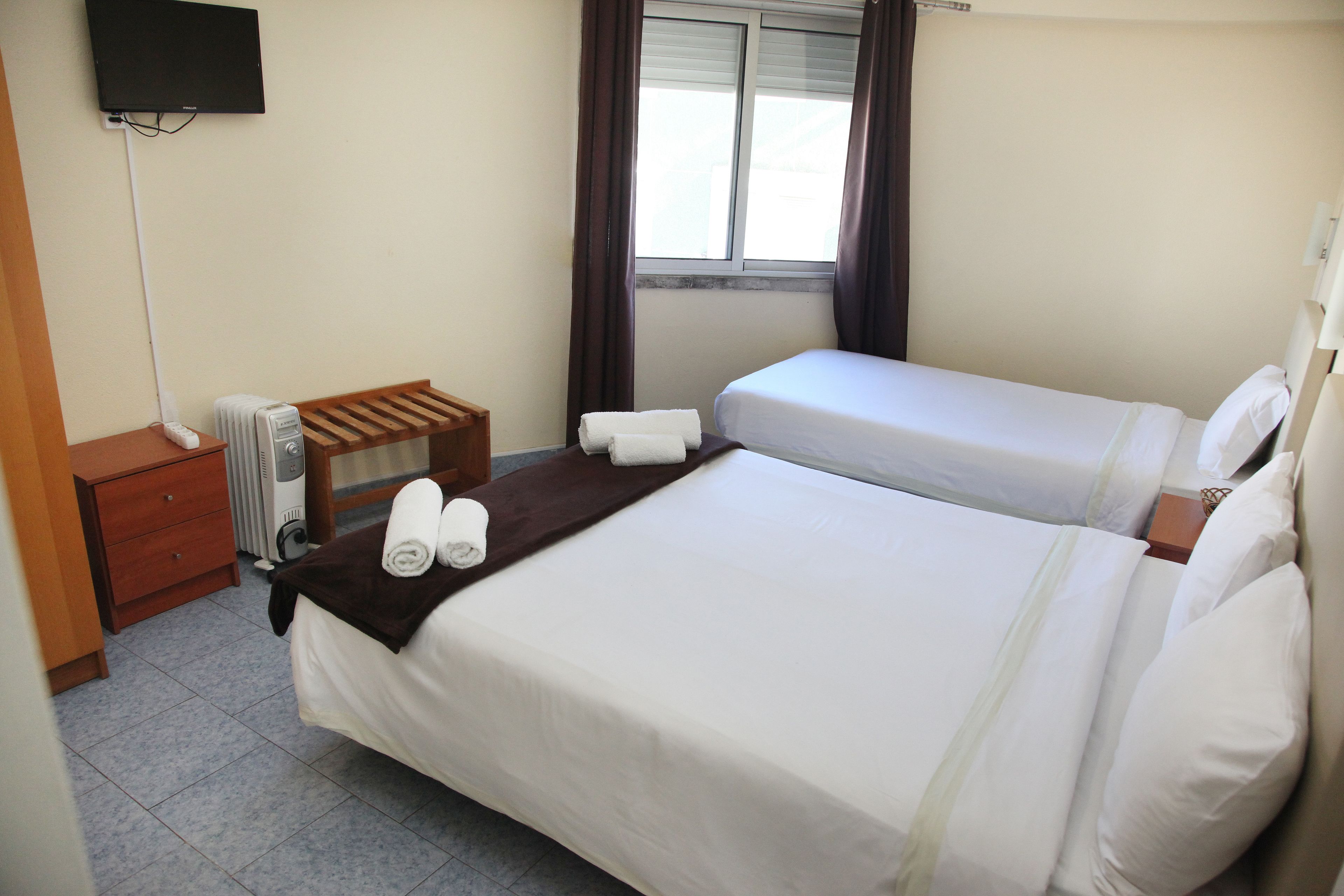 Ein Hotelzimmer mit zwei Betten, einem an der Wand befestigten Fernseher, einem Heizgerät und einem Fenster mit Vorhängen.