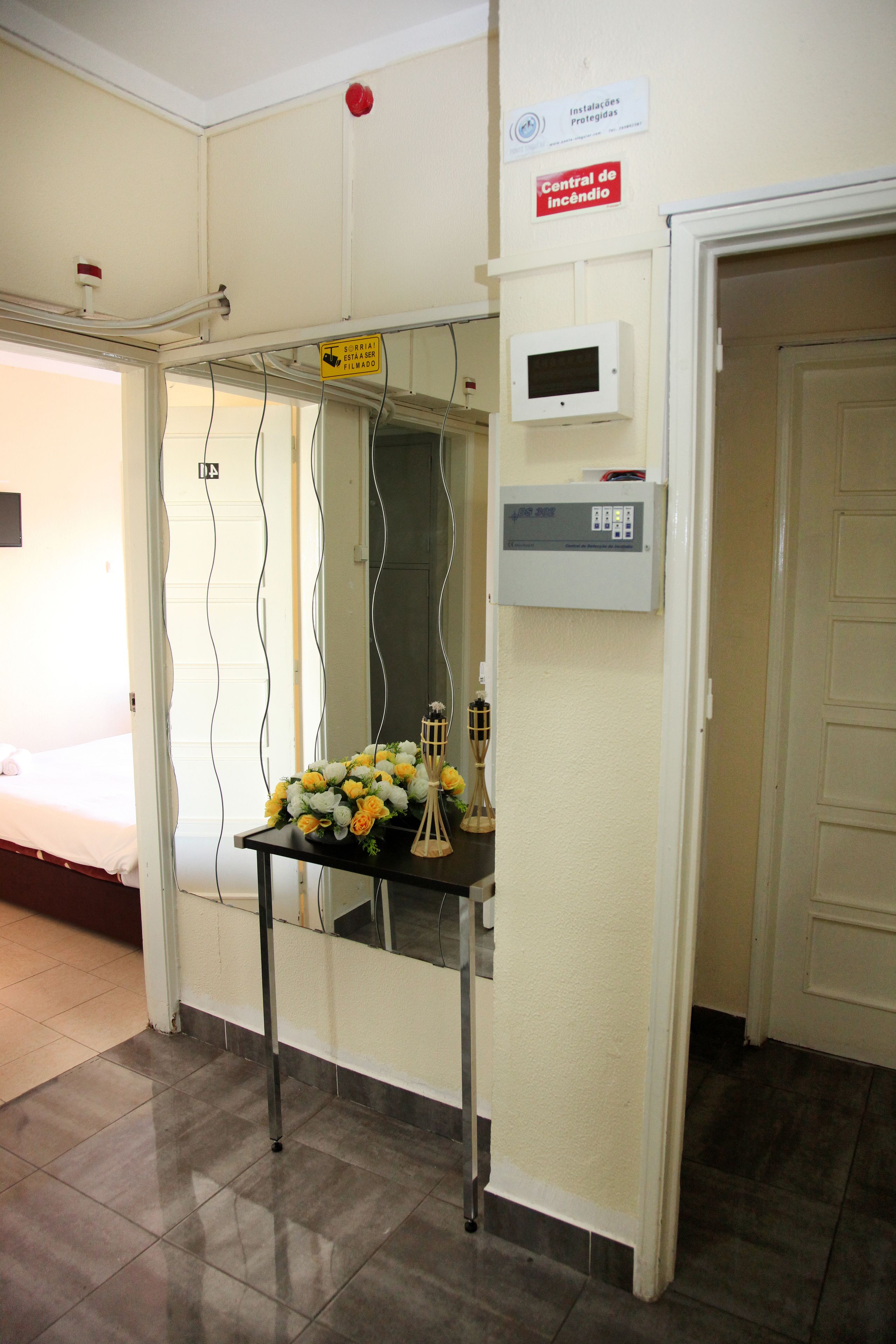 A hotel room with a bed, a table with flowers, a fire alarm, and a control panel.