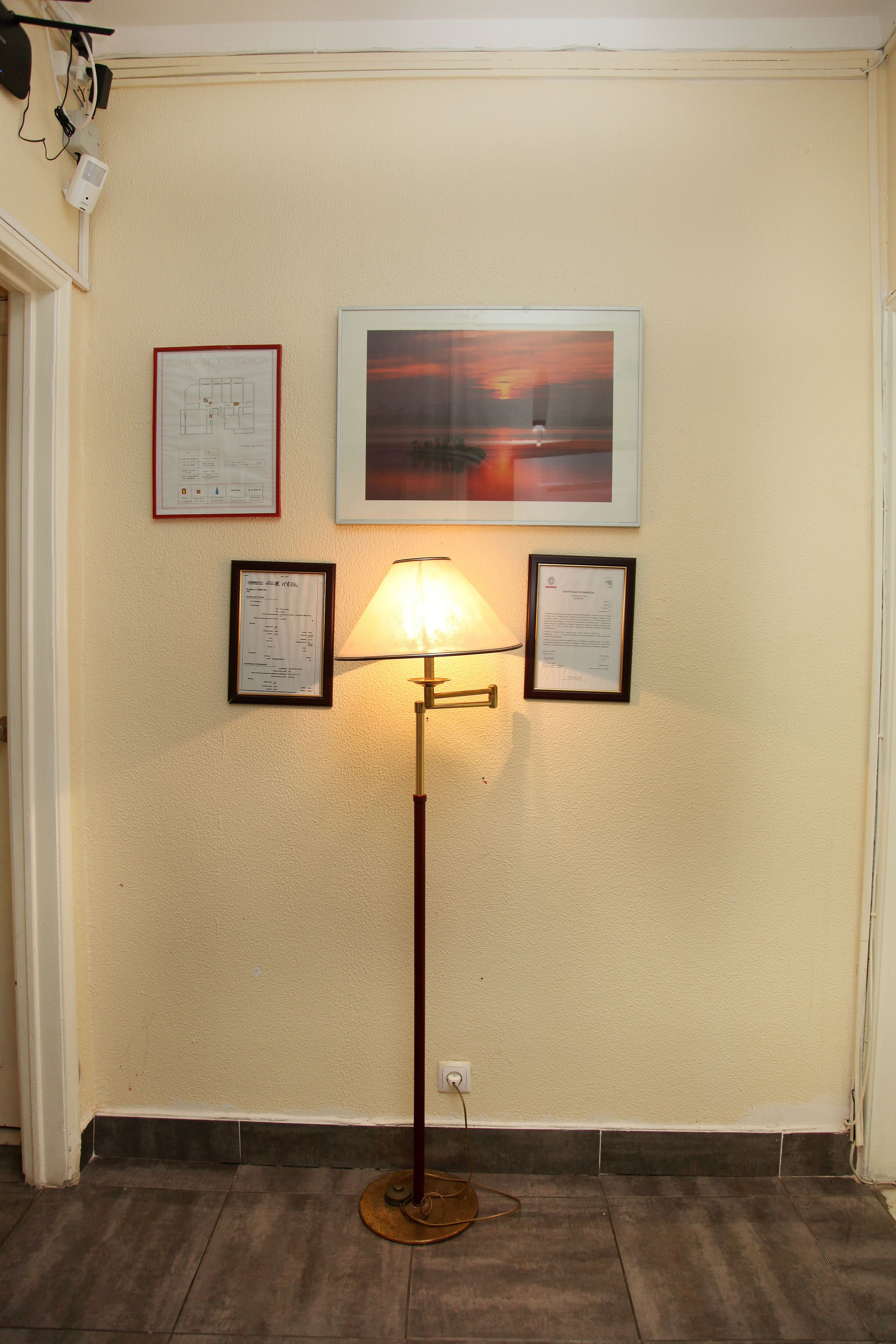 A hallway with a floor lamp, framed certificates, and a sunset painting on the wall.