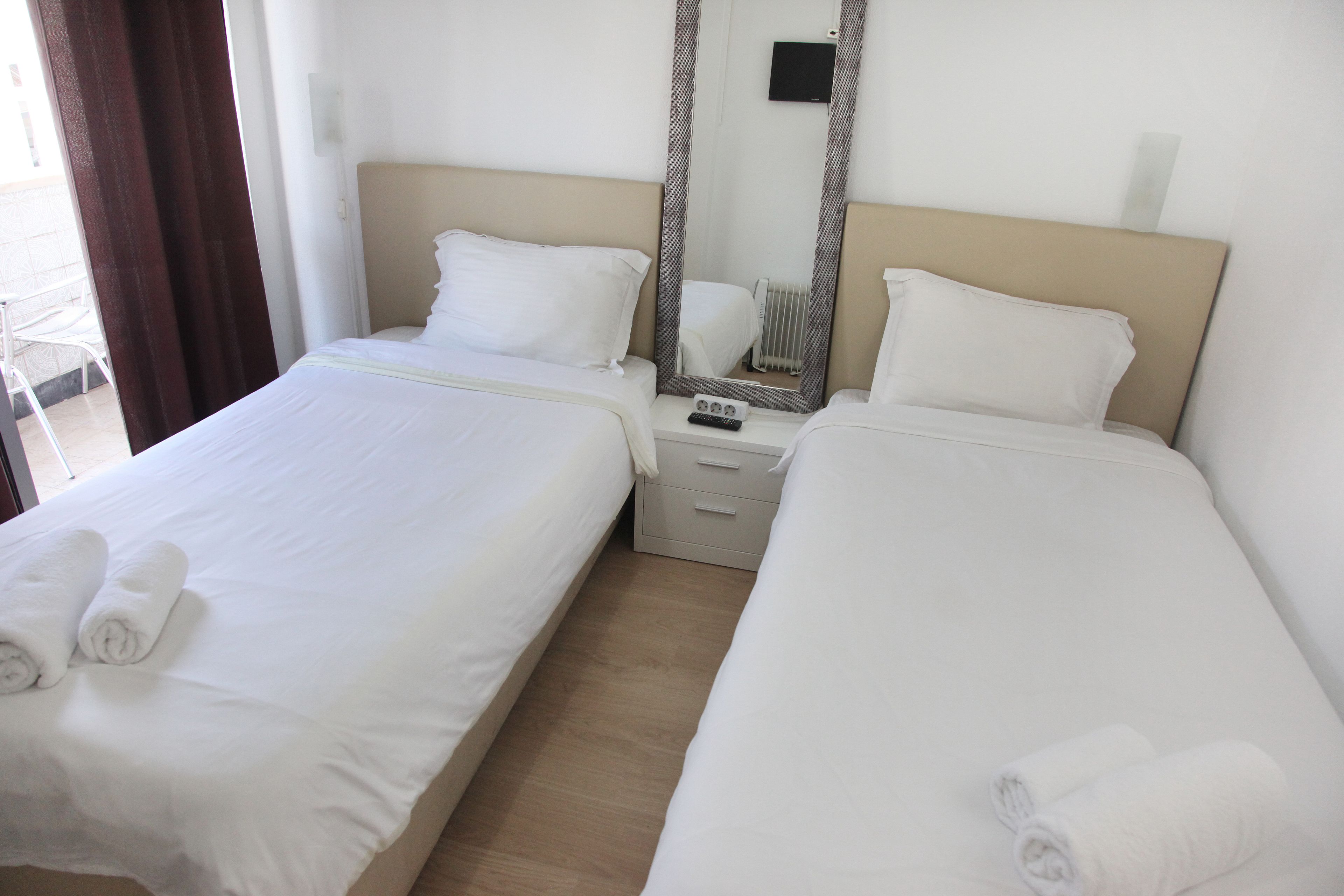 A hotel room with two beds, each with white linens and rolled towels. There is a bedside table with a remote control and a mirror above it.