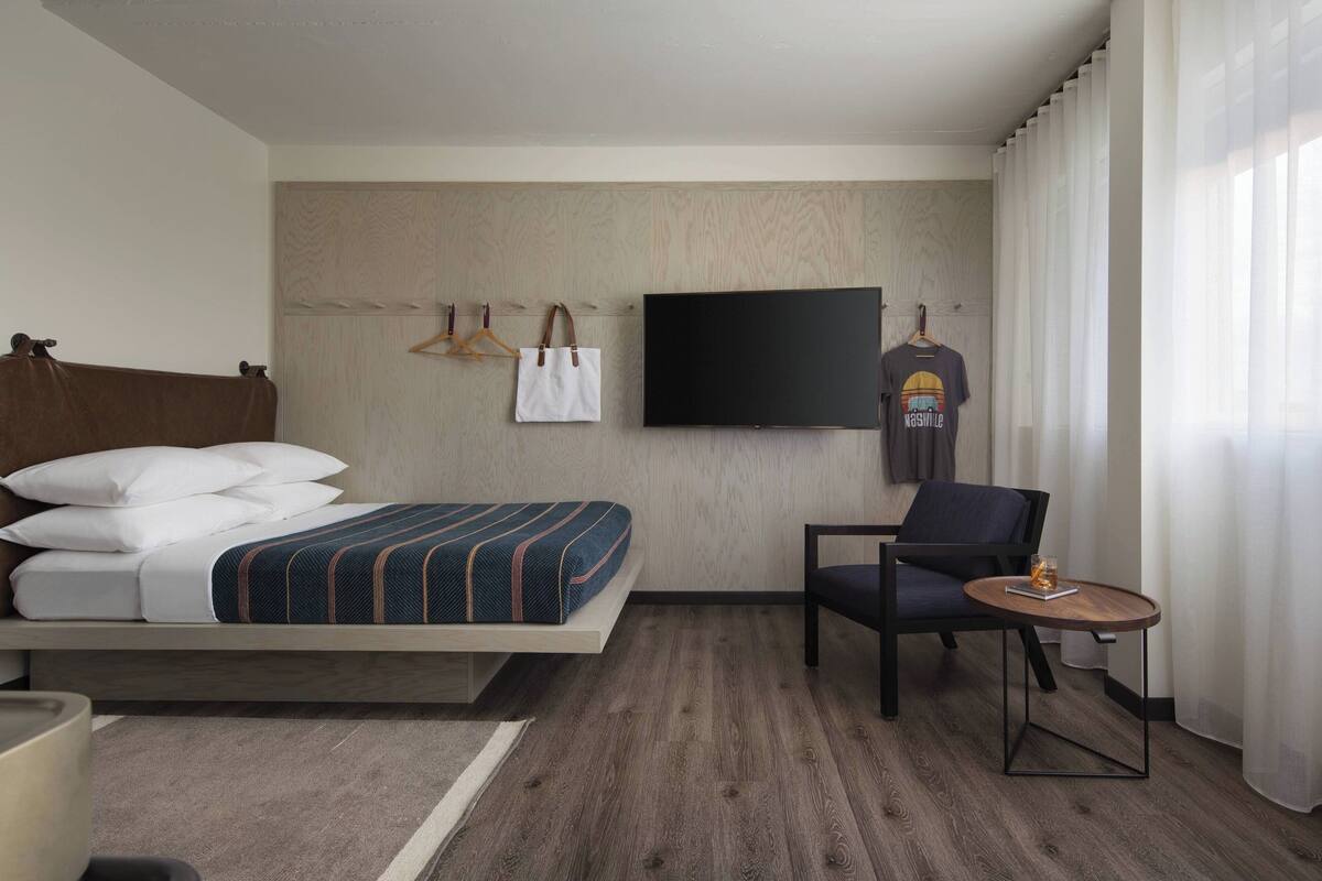 Ein Hotelzimmer mit Bett, Sessel, kleinem Tisch, Fernseher und Kleiderhaken mit Kleiderbügeln.