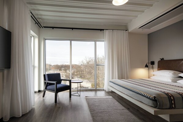Una habitación de hotel moderna con una cama grande, una silla, una mesita y vistas a la ciudad a través de amplios ventanales.