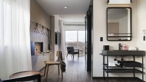Habitación de hotel moderna con suelo de madera, un espejo grande, un lavamanos negro y vistas de la ciudad a través de amplios ventanales.