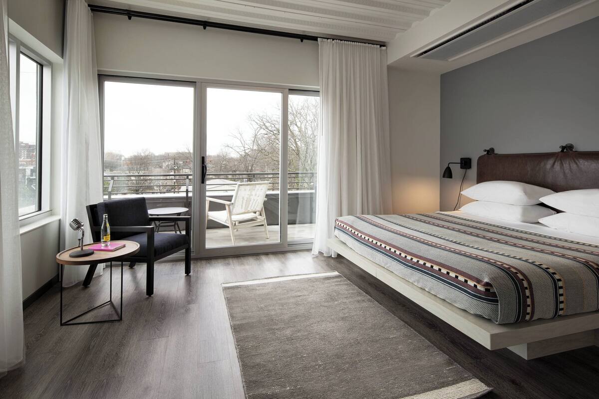 Ein modernes Hotelzimmer mit einem großen Bett, einem kleinen Tisch mit Stuhl, einem Balkon mit Blick auf die Stadt und einem großen Fenster mit Vorhängen.