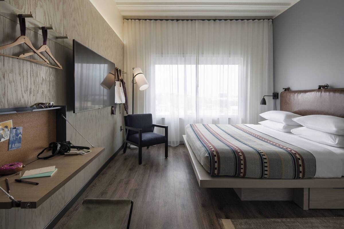Ein Hotelzimmer mit Bett, Schreibtisch mit Stuhl, einem am Wand montierten Flachbildfernseher, einer Lampe und einem Fenster mit transparenten Vorhängen.