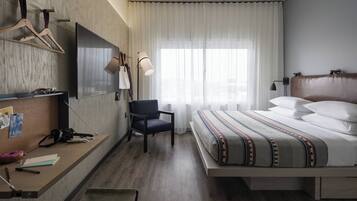 Habitación de hotel con cama, escritorio con silla, televisor de pantalla plana montado en la pared, lámpara y ventana con cortinas translúcidas.