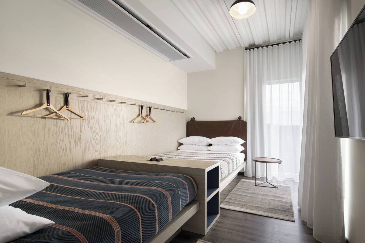 Ein modernes Hotelzimmer mit einem Holz-Kopfteil, zwei Betten mit gestreifter Bettwäsche, einem Nachttisch, einem kleinen runden Tisch und einem großen Fenster mit transparenten Vorhängen.