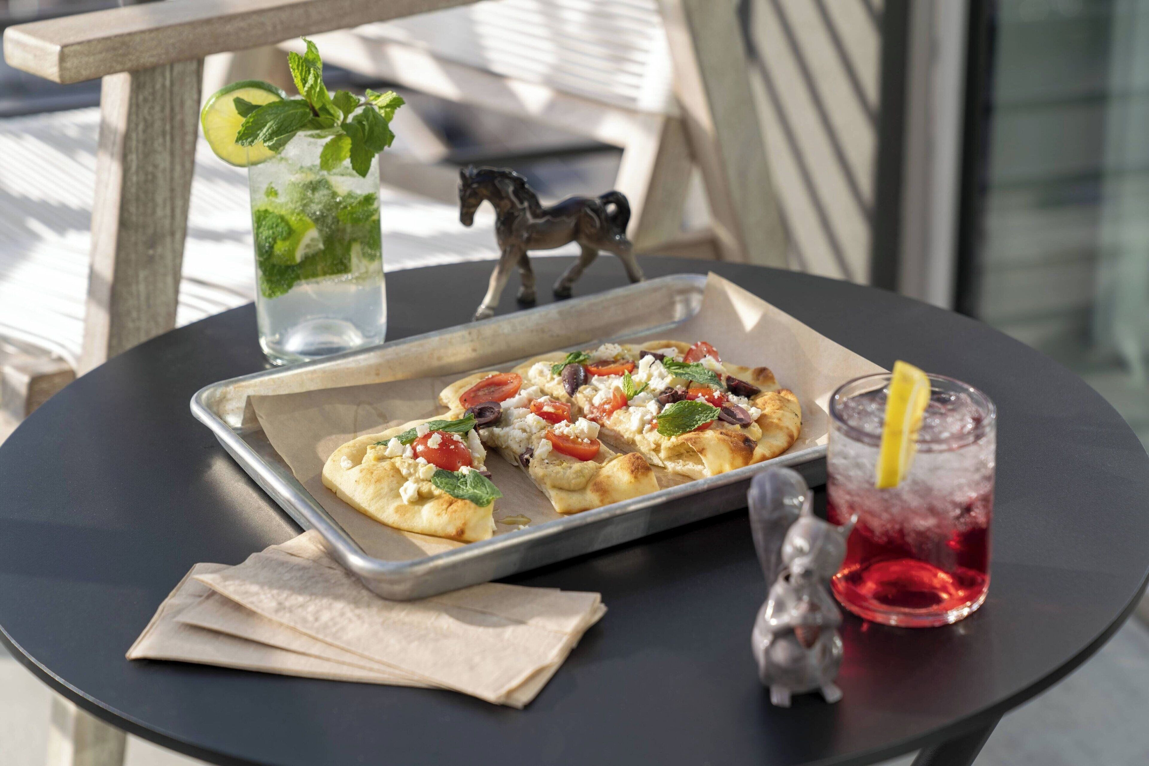 Una bandeja con porciones de pizza sobre una mesa, un vaso de bebida roja con una rodaja de limón y una ramita de menta.