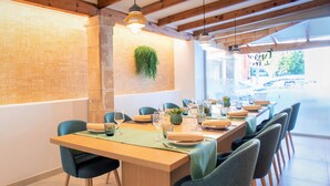 Un coin repas est aménagé autour d’une longue table en bois, dressée pour six personnes, et entourée de six chaises. Une plante suspendue décore le mur, et la pièce présente un design moderne et épuré, avec des poutres en bois apparentes au plafond.