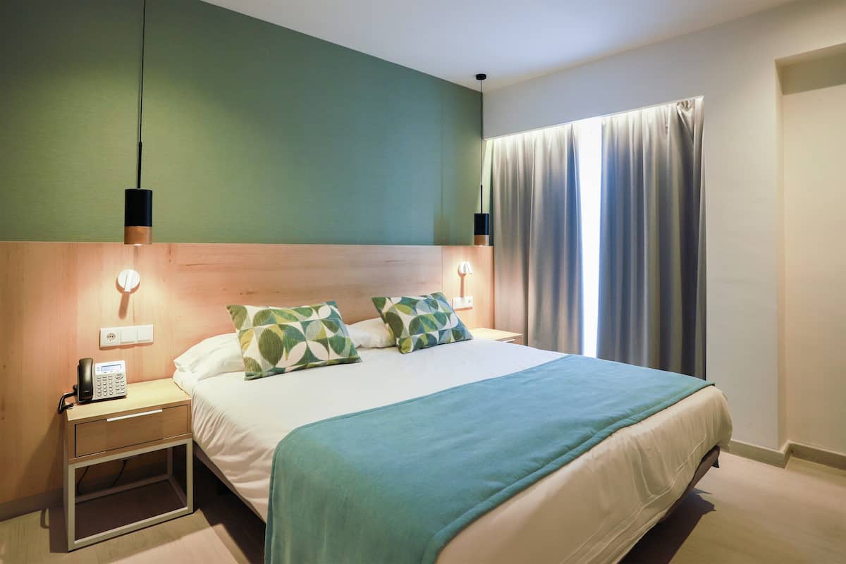 Una cama bien hecha con una colcha verde, dos almohadas estampadas, una lámpara de noche y un teléfono sobre una mesita de madera.