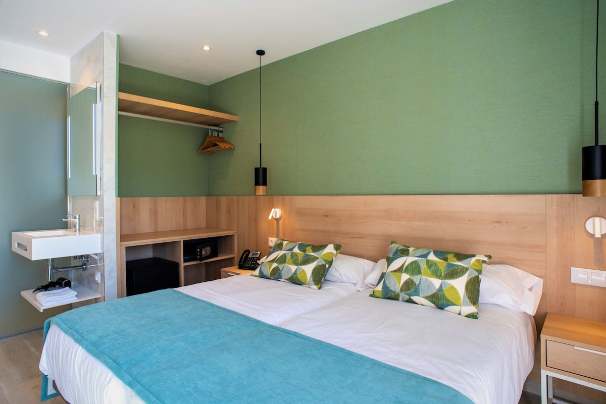 Habitación de hotel con cama, mesita de noche, espejo, estante con abrigo y lámpara colgante.