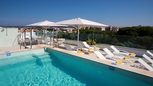 Une piscine d’un bleu éclatant, bordée de chaises longues et d’ombrelles, entourée d’une barrière de verre, et offrant une vue sur les bâtiments en arrière-plan.