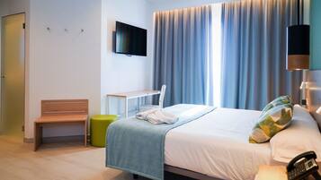 A hotel room with a bed, a bedside table with a phone, a small desk, a chair, a bench, and a TV on the wall.