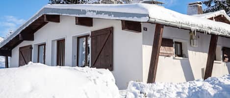 A snow-covered building with a sloped roof, wooden doors, and a chimney.