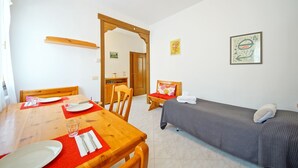 A room with a wooden dining table set with plates and glasses, a single bed with a gray blanket, and two wall-mounted artworks.