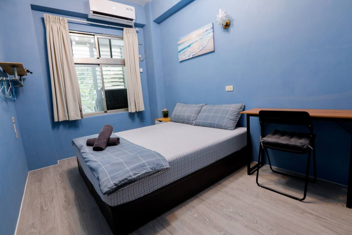 A compact bedroom with a bed, a small table, a chair, and a window with curtains.