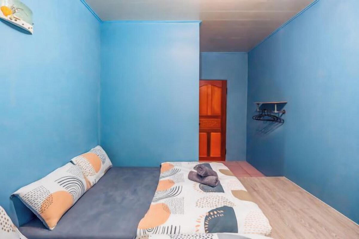 A room with blue walls, a bed with patterned bedding, a wooden floor, and a door in the background.