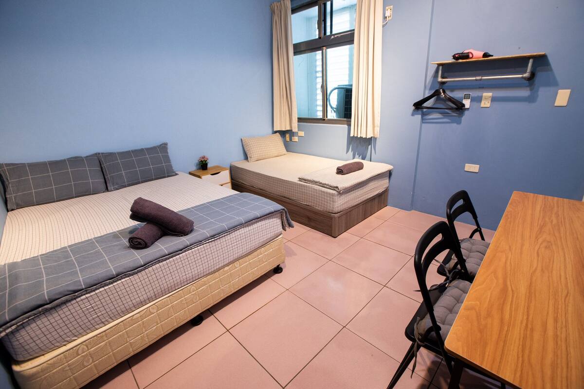 A bedroom with two beds, a wooden table, a chair, a window with curtains, and a wall-mounted shelf with a heater.
