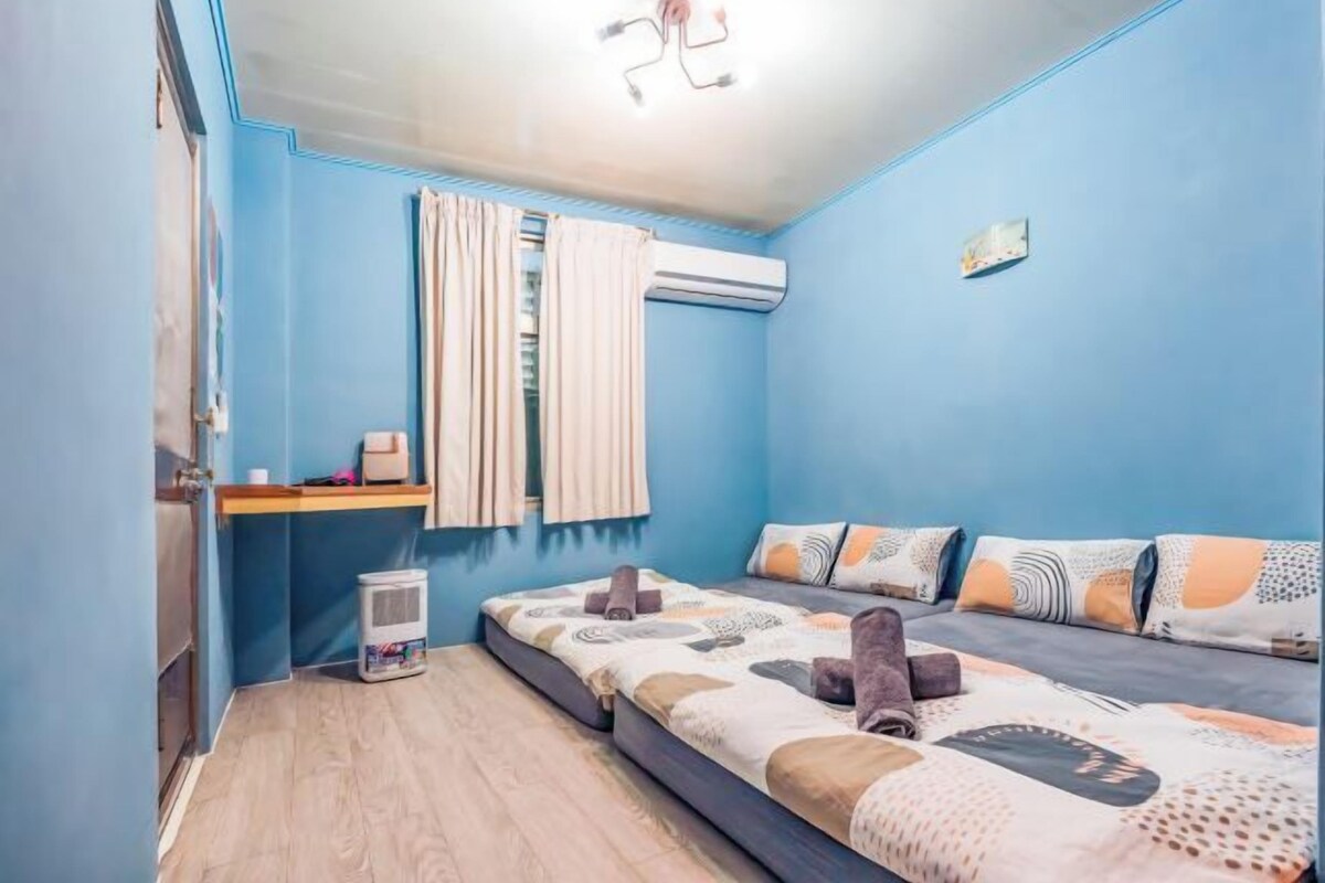 A bedroom with a bed, a small table, an air conditioning unit, and a window with curtains.