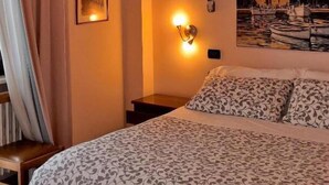 Ein Schlafzimmer mit einem Bett, einem Nachttisch, einer Wandlampe, einem Gemälde und einem Fenster mit Vorhängen.