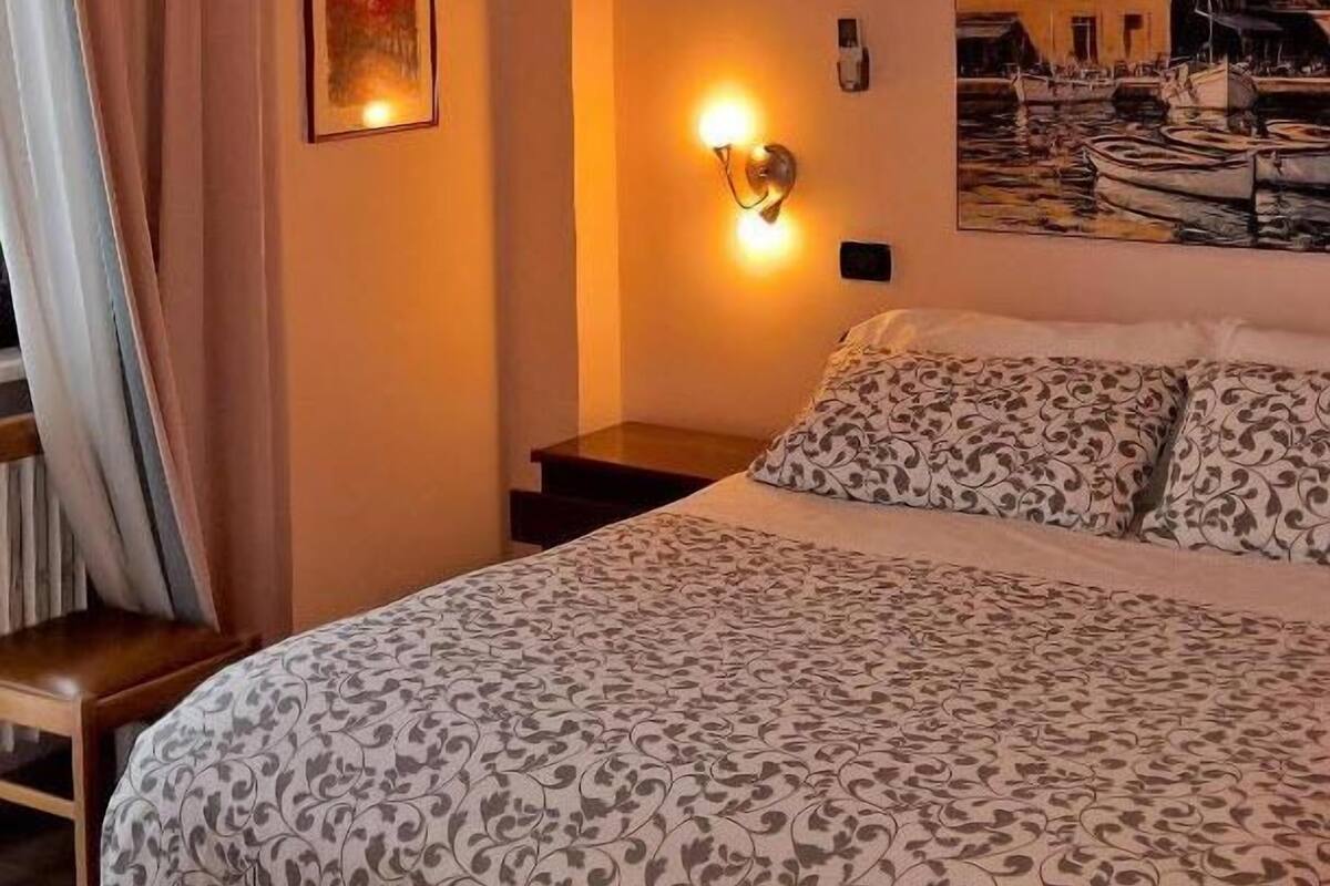 A bedroom with a bed, a nightstand, a wall-mounted lamp, a painting, and a window with curtains.