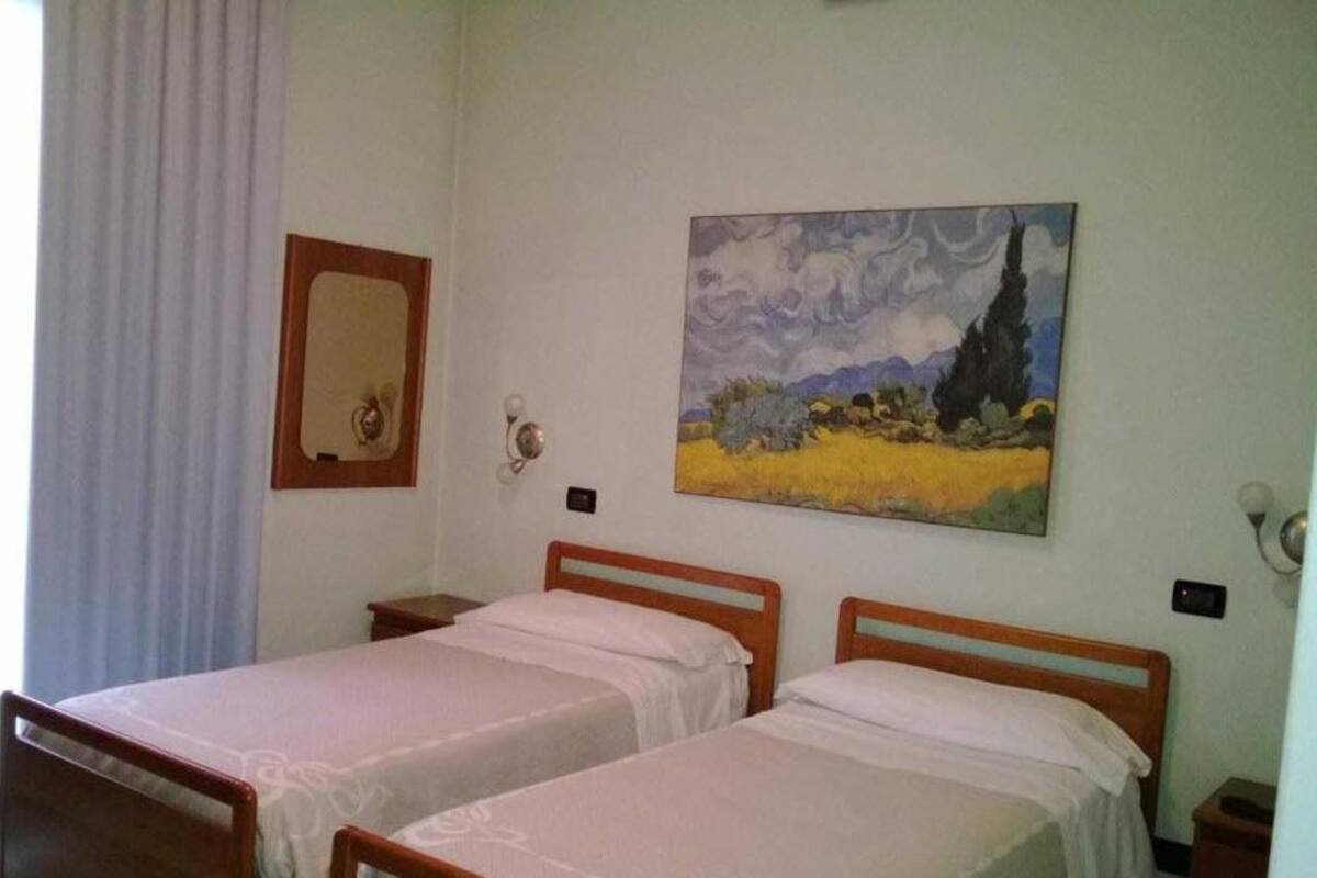 A room with two single beds, a painting on the wall, a mirror, and a window with curtains.
