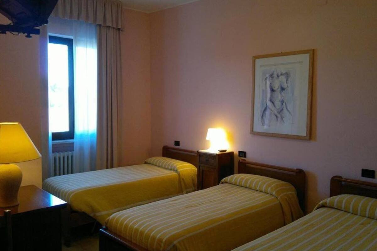 A hotel room with two single beds, a nightstand with a lamp, a window with curtains, and a framed artwork on the wall.