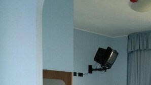 Ein Zimmer mit einem Bett, einem Holzschrank, einem kleinen Tisch, einem Spiegel und einem an der Wand befestigten Fernseher.