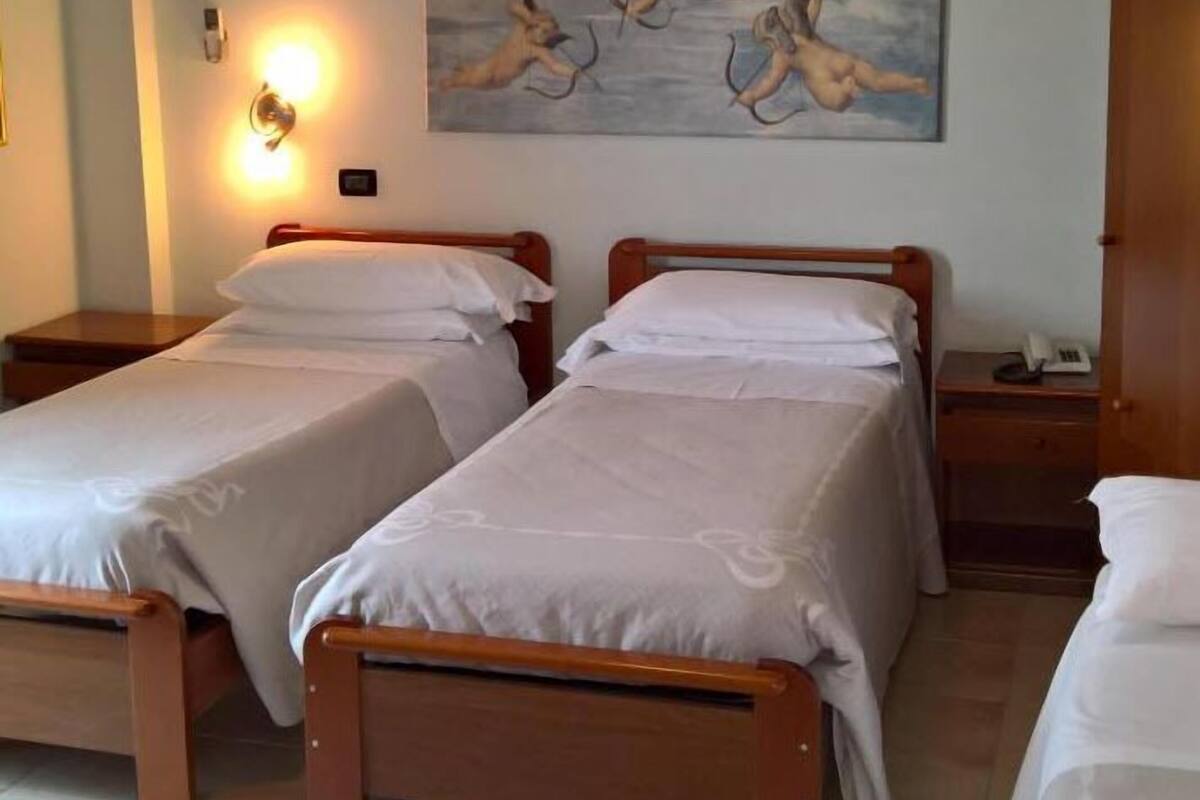 A hotel room with two wooden beds, white bedding, a nightstand with a lamp, and a painting of cherubs on the wall.