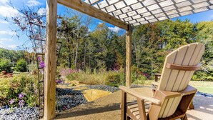 Una pérgola de madera con techo a cuadros, una silla de madera, y un jardín con senderos de grava y diversas plantas.