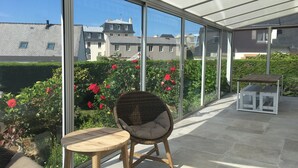 En täckt uteplats med en flätstol och ett litet bord, stora fönster som erbjuder utsikt över byggnader och en trädgård med röda blommor.