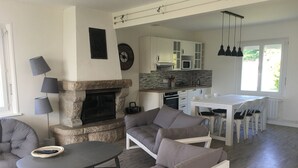 Ett modernt vardagsrum med en stenbeklädd öppen spis, grå soffor, en matplats med ett vitt bord och stolar samt ett kök med vita skåp och en mikrovågsugn.